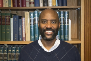 Headshot of Curtis Small, a bald smiling man with black facial hair, wearing a blue sweater with a white undershirt, standing in front of a bookshelf