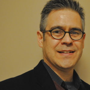 headshot of R. Darrell Meadows, a man with short gray hair and black framed glasses, wearing a black suit jacket with a blue shirt, in front of a yellow background.