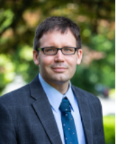 Headshot image of Kevin Wisniewski. A man with short black hair wearing dark framed glasses, dressed in a blue suit with a blue shirt and dark blue tie.