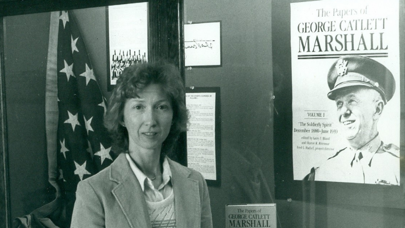 Black and white photo of Sharon Ritenour Stevens, standing in front of a museum exhibit of the papers of George Catlett Marshall and an American Flag