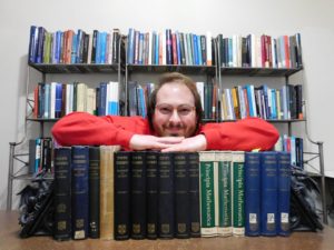 Landon D. Elkin poses with his arms crossed on multiple editions of Whitehead & Russell's Principia Mathematica, in front of several bookshelves